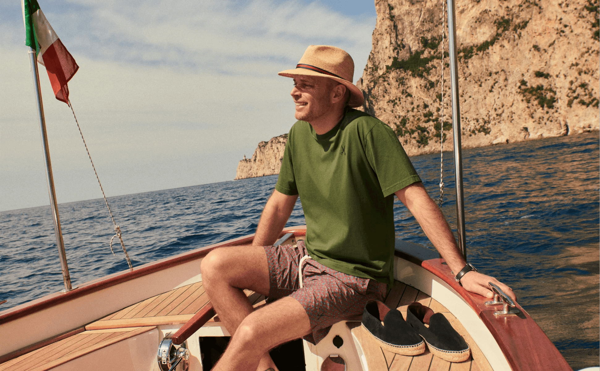 A man seated on a wooden boat beside black espadrilles with a rocky coastline behind him.